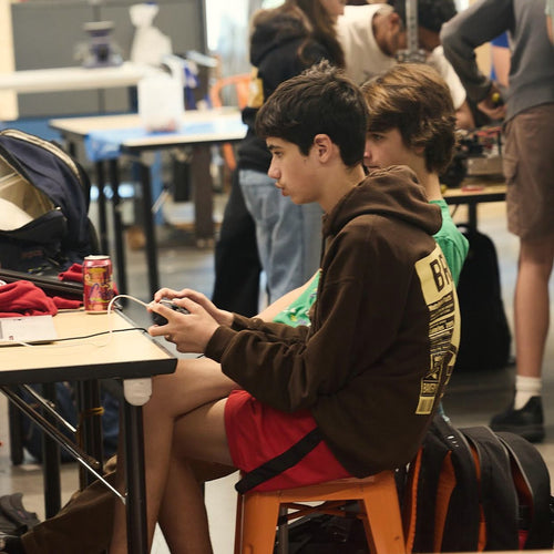 A middle schooler works on a device at a makerspace table while classmates build behind him.