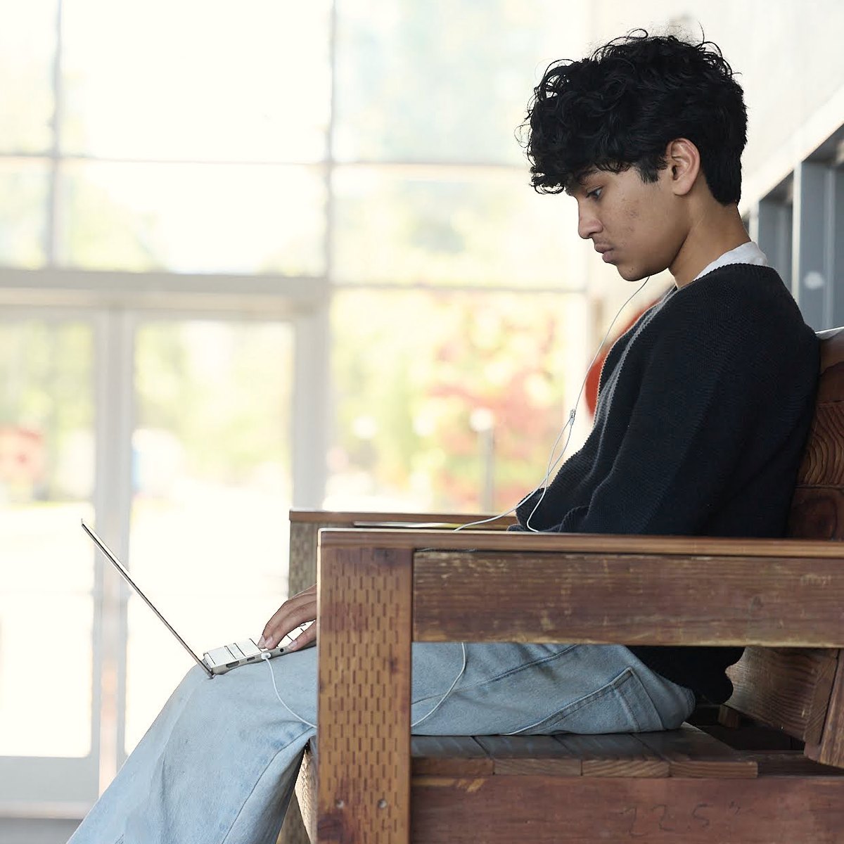 A student works on a laptop on a wooden bench beside tall, sunlit windows.