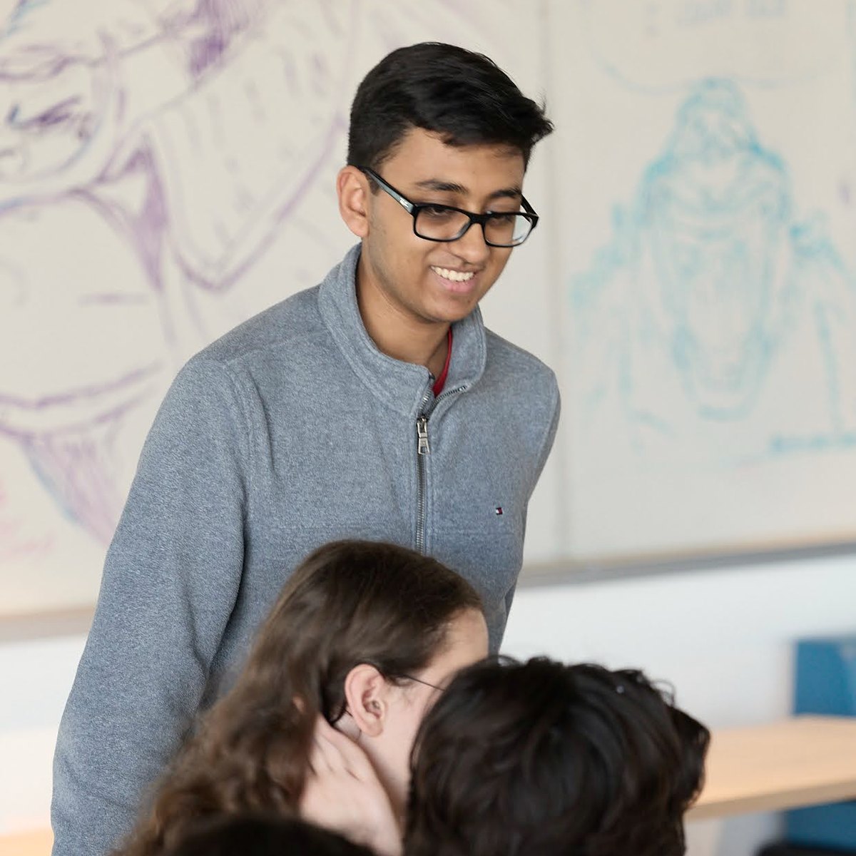 A mentor smiles down at students working at a table, whiteboard sketches behind him.